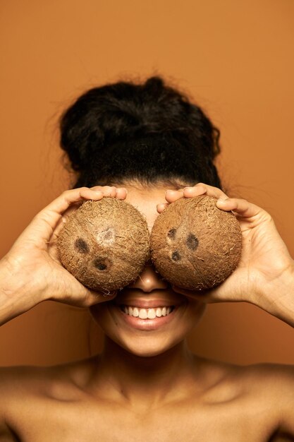 Mujer con piel perfecta y cabello en un moño escondiendo sus ojos detrás de dos cocos, posando aislado en naranja