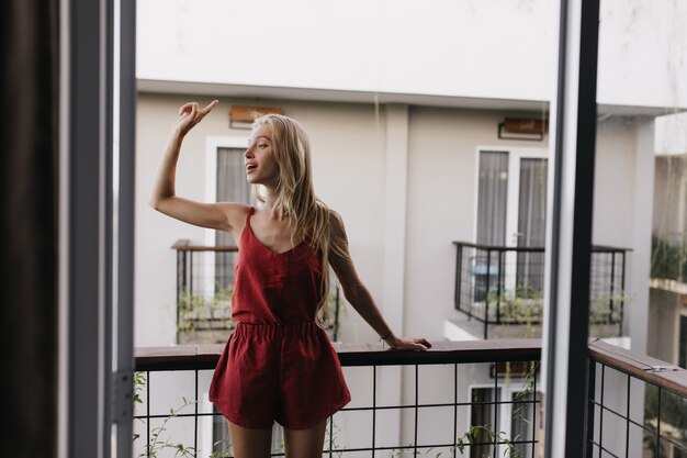 mujer en pijama de pie en el balcón y mirando a su alrededor. Modelo de mujer emocionada con largo cabello rubio disfrutando de la mañana.
