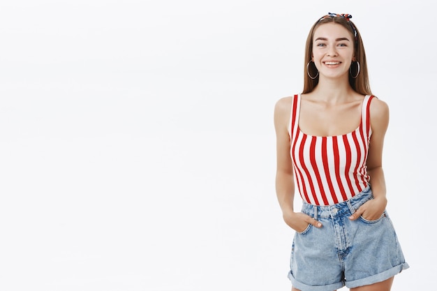 Mujer pinup caliente en la parte superior de rayas de moda y pantalones cortos con diadema tomados de la mano en los bolsillos sonriendo con alegría posando