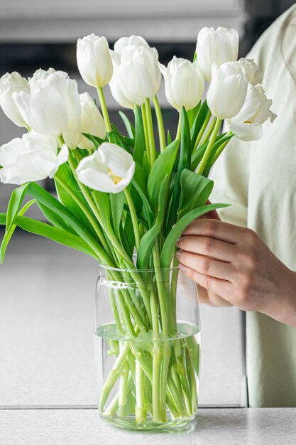 Mujer poniendo un ramo de flores de tulipán blanco en un jarrón en la cocina