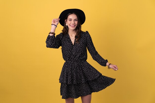 Mujer posando aislada en amarillo vistiendo un vestido negro punteado y un sombrero negro con estilo primavera verano accesorios de estilo de moda, sonriendo feliz humor divirtiéndose