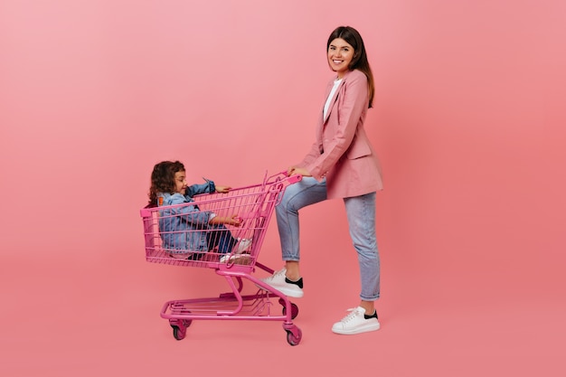 Mujer posando con hija después de ir de compras. Niña preadolescente despreocupada sentada en el carro de la tienda.