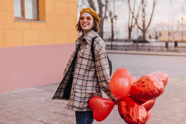 Mujer positiva en abrigo a cuadros sonriendo contra la pared de la ciudad