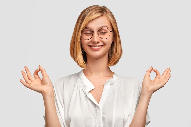 La mujer positiva alegre hace un buen gesto con ambas manos, tiene un corte de pelo moderno, cierra los ojos, vestida con camisa blanca