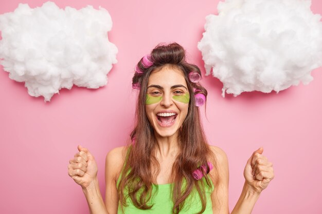 La mujer positiva aprieta los puños celebra algo aplica rulos parches de belleza verde debajo de los ojos quiere tener un gran corte de pelo mañana se trata a sí misma aislada en la pared rosa