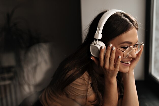 Mujer positiva en auriculares enormes blancos se pone gafas y sonríe, apoyándose en el alféizar de la ventana negra.