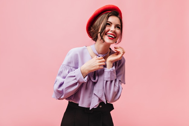 Mujer positiva en boina roja y blusa lila se ríe sobre fondo rosa.