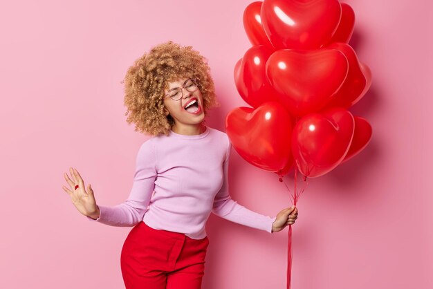 Una mujer positiva de cabello rizado con maquillaje filtrado canta una canción que mantiene la boca abierta, usa un suéter casual y pantalones con un montón de globos de corazón disfruta de un evento especial aislado sobre un fondo rosado