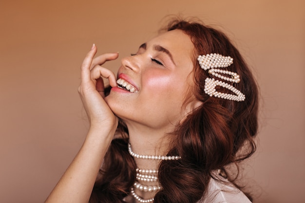 Foto gratuita mujer positiva con cabello rizado se ríe y se muerde el dedo. retrato de mujer con horquillas blancas y collar de perlas.