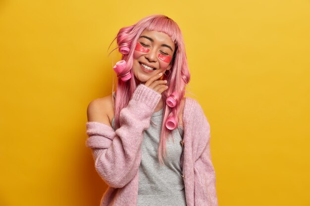 Mujer positiva con cabello rosado, inclina la cabeza, toca la cara suavemente, disfruta de una piel suave, tiene una sonrisa con dientes, hace un peinado, se preocupa por la piel debajo de los ojos