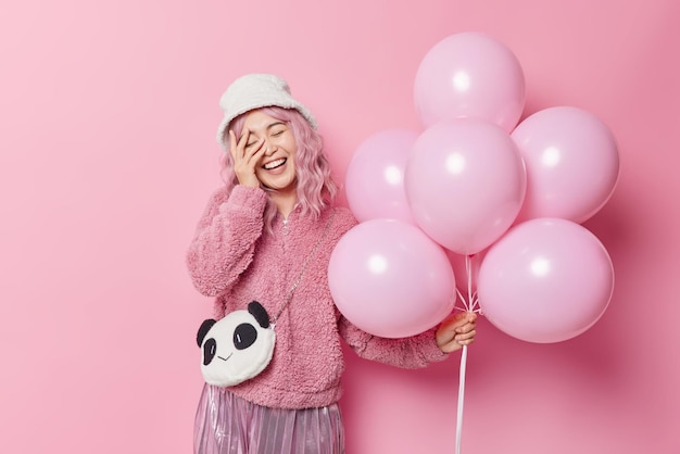 La mujer positiva con cabello rosado mantiene la mano en la cara sonríe alegremente vestida con ropa de abrigo se divierte en ocasiones especiales disfruta de la celebración sostiene un montón de globos inflados aislados sobre una pared rosa