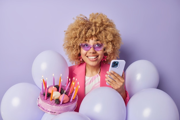 La mujer positiva celebra el aniversario vestida con un elegante teléfono móvil que sostiene la ropa y recibe un mensaje de poses de felicitación con un sabroso pastel de cumpleaños alrededor de globos inflados aislados sobre una pared púrpura