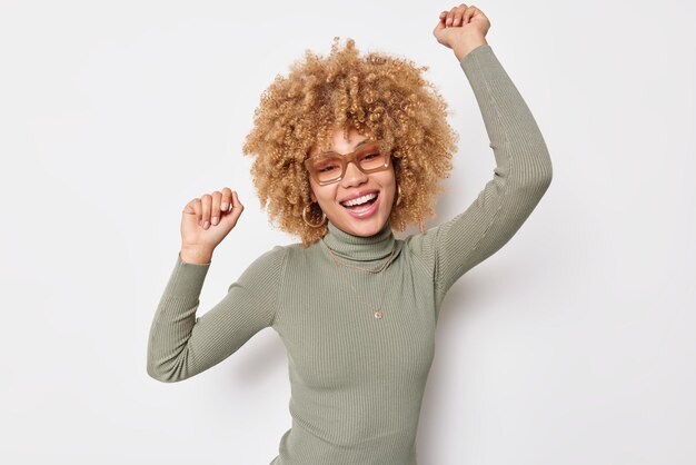 La mujer positiva y despreocupada de buen humor baila con los brazos levantados, se siente feliz, se mueve activamente contra el fondo blanco, usa gafas y el cuello de tortuga se divierte. Chica de pelo rizado relajado interior