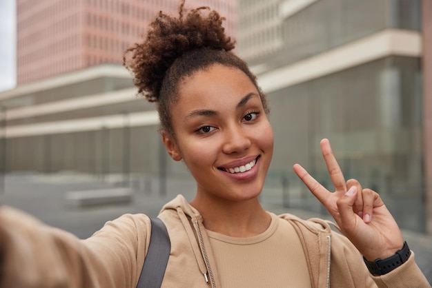 Una mujer positiva y despreocupada con el pelo rizado vestido con capucha sonríe felizmente hace un gesto de paz y se toma selfie en un lugar urbano estando de buen humor después del entrenamiento deportivo Emociones de la gente y estilo de vida deportivo