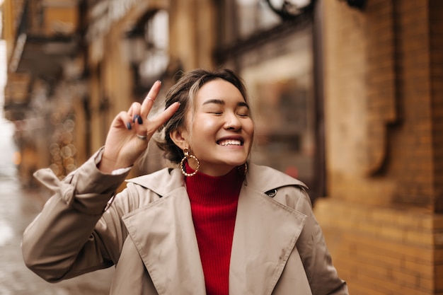 Mujer positiva en enormes pendientes riendo con los ojos cerrados contra la pared del edificio