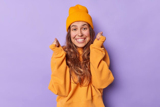 Una mujer positiva eufórica aprieta los puños esperando resultados importantes sonríe ampliamente tiene dientes blancos se siente emocionada se siente entusiasta y optimista usa un jersey suelto informal y poses de sombrero en el interior.