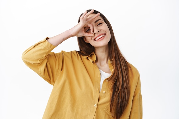 Mujer positiva y feliz haciendo el signo de OK en el ojo y sonriendo mostrando aprobación como y de acuerdo decir que sí hacer el gesto de estar bien complacido con el buen resultado de pie sobre fondo blanco