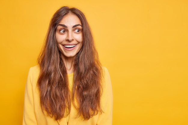 La mujer positiva feliz con el pelo largo oscuro sonríe agradablemente enfocada a un lado tiene modelos de expresión alegre curiosa contra el espacio de copia en blanco de la pared amarilla vívida para su información. Concepto de emociones