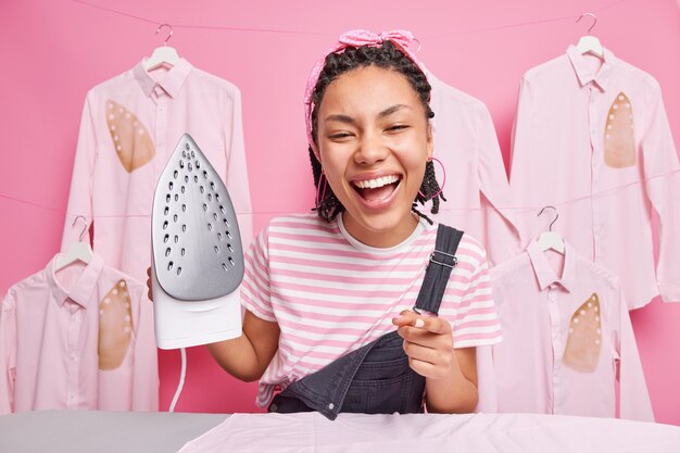 La mujer positiva feliz trabaja alrededor de los puntos de la casa directamente en ti sonríe ampliamente sostiene la plancha eléctrica yendo a lavar la ropa estando de buen humor aislado sobre una pared rosa