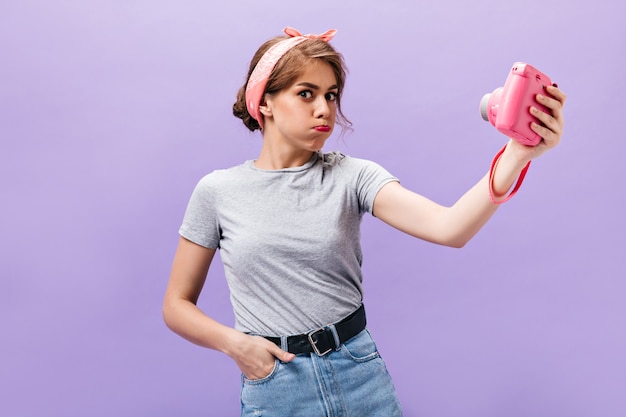 La mujer positiva hace una cara graciosa y sostiene una cámara rosa. Chica joven fresca en camiseta gris de moda y piel de mezclilla con cinturón negro posando.