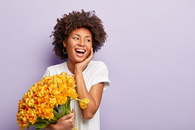 La mujer positiva interior se ríe y se divierte, disfruta del aroma de los tulipanes amarillos de primavera, usa una camiseta blanca informal, aislada sobre una pared lila, espacio en blanco para su contenido promocional. Tiempo de primavera