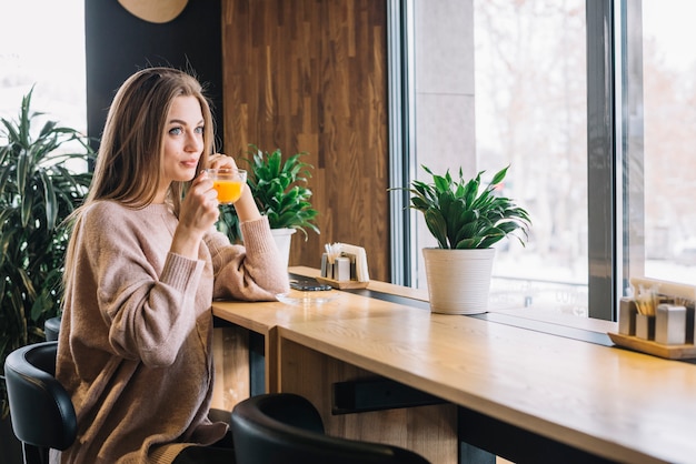 Mujer positiva joven elegante que sostiene la taza de bebida en el contador de la barra cerca de ventana en café