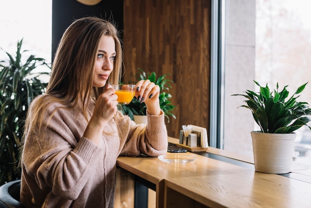 Mujer positiva joven elegante que sostiene la taza de bebida en el contador de la barra cerca de ventana en café