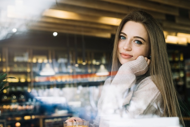 Foto gratuita mujer positiva joven elegante con la taza de bebida en café a través de la ventana