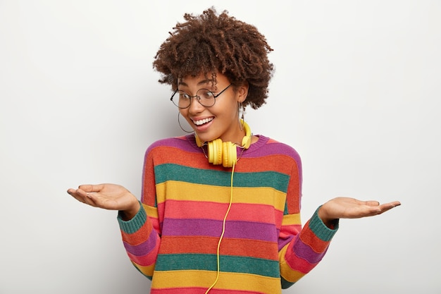 Mujer positiva levanta palmas, sostiene dos objetos invisibles, toma decisiones entre buenas propuestas, usa lentes ópticos y suéter de rayas