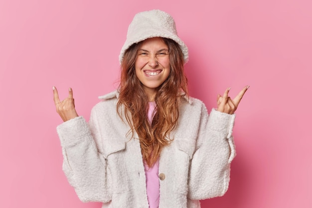 Foto gratuita la mujer positiva con el pelo largo muerde los labios hace que el gesto del heavy metal del rock n roll disfruta de la música fresca estando de buen humor viste un panamá blanco y un abrigo aislado sobre fondo rosa. concepto de lenguaje corporal