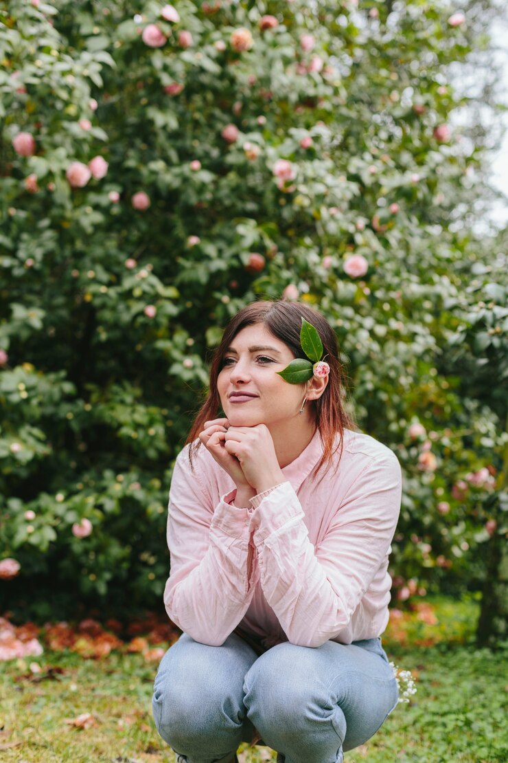 Mujer positiva pensativa con planta en cabello cerca de flores rosadas que crecen en ramitas verdes