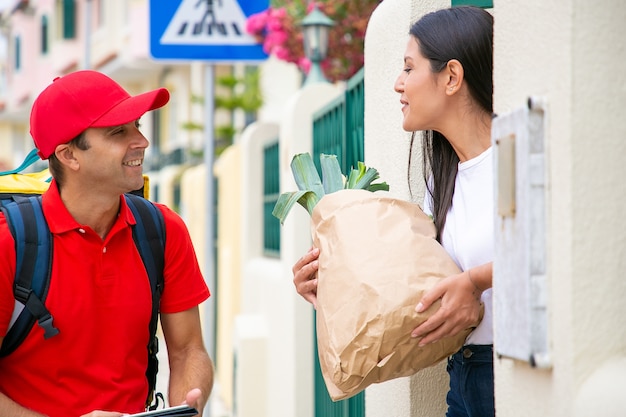 Mujer positiva que recibe comida de la tienda de comestibles, sosteniendo el paquete de papel con anuncios de verduras verdes agradeciendo al mensajero en uniforme rojo. Concepto de servicio de envío o entrega