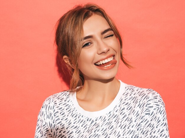 Mujer positiva sonriendo. Modelo divertido posando junto a la pared rosa en el estudio. Muestra lengua y guiños