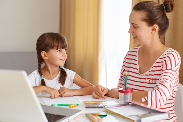 Mujer positiva con su hija posando en la sala de estar en la mesa, madre ayudando a su hija con lecciones, explicando la nueva regla, educación a distancia en línea.