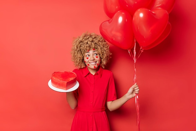 Foto gratuita la mujer positiva tiene el cabello rubio rizado, usa un vestido festivo, sostiene un montón de globos de corazón y un pastel dulce se prepara para las vacaciones aislado sobre un fondo rojo vivo concepto de personas y celebraciones