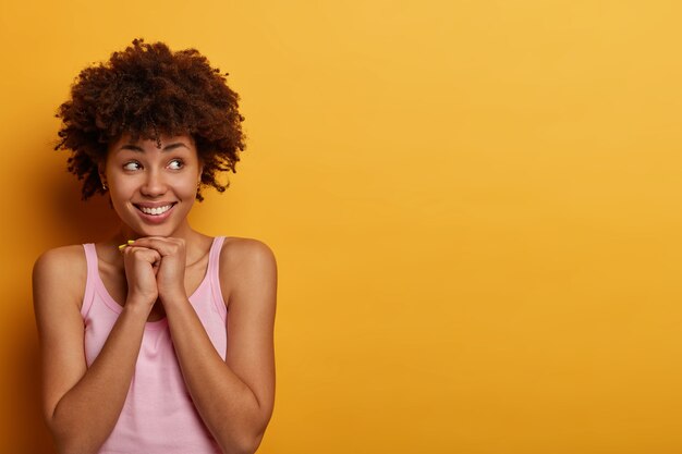 La mujer positiva tiene una expresión de satisfacción pensativa, mantiene las manos debajo de la barbilla, mira a un lado, se viste de manera informal, piensa en planes futuros, aislada sobre una pared amarilla, espacio en blanco para su texto