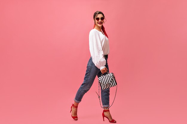 Mujer positiva en traje elegante se mueve sobre fondo rosa. Mujer bonita en blusa blanca y tacones rojos está sonriendo a la cámara.