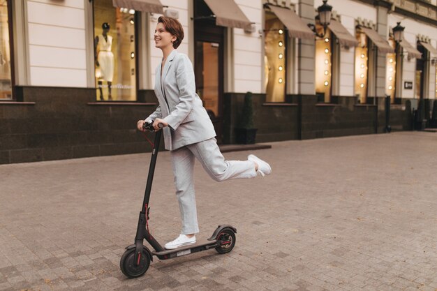 Mujer positiva en traje gris montando scooter al aire libre. Encantadora chica de pelo corto en chaqueta y pantalón sonriendo y caminando por la ciudad