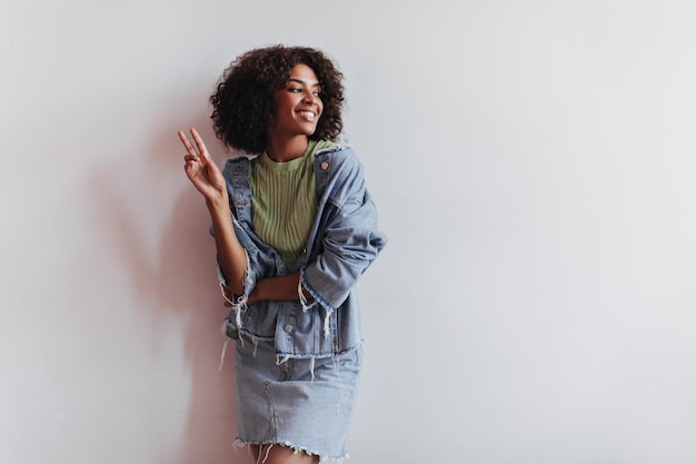 Foto gratuita mujer positiva en traje de mezclilla se apoya en la pared blanca y muestra el signo de la paz retrato de chica elegante en falda azul y chaqueta sobre fondo aislado