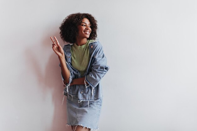 Mujer positiva en traje de mezclilla se apoya en la pared blanca y muestra el signo de la paz Retrato de chica elegante en falda azul y chaqueta sobre fondo aislado