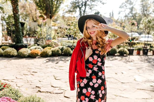 Mujer positiva en vestido negro con estampado floral, chaqueta de gamuza y sombrero muestra el signo de la paz y posa en el jardín.