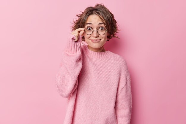 La mujer positivamente sorprendida mantiene la mano en el borde de las gafas, sonríe, reacciona agradablemente ante las buenas noticias, usa un puente casual de cachemira, escucha algo impresionante aislado sobre un fondo rosa. Reacción y emociones