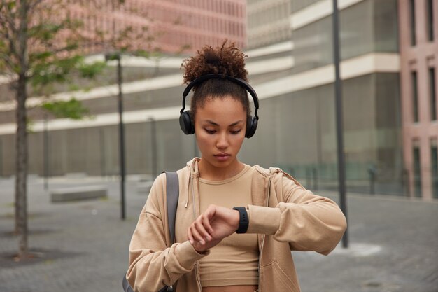 La mujer practica deporte monitorea regularmente sus resultados de acondicionamiento físico en un reloj inteligente, escucha música mientras se entrena vestida con poses de ropa deportiva en un entorno urbano