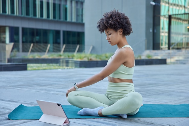 Mujer practica yoga se sienta con las piernas cruzadas en la estera de fitness mira videos a través de modernas poses de tableta portátil afuera