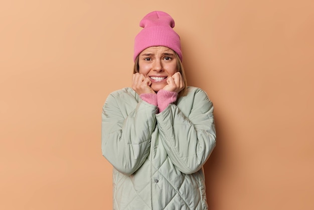 La mujer preocupada y estresada aprieta los dientes se ve ansiosa y preocupada por los miedos de algo aterrador tiembla por el miedo usa sombrero y chaqueta aislados sobre fondo beige Reacciones de emociones negativas