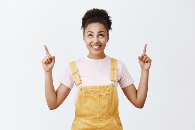 Mujer preparándose para subir al escenario, dándole la dirección de Dios para ayudarla. Encantadora mujer joven feliz y despreocupada con piel oscura con un mono amarillo, mirando y apuntando hacia arriba con los dedos índices, mirando al cielo