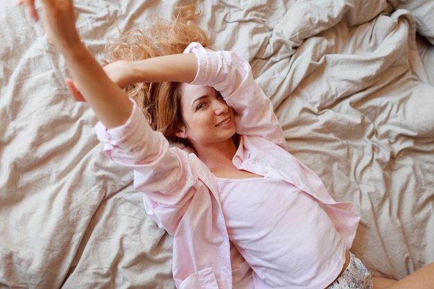 Mujer principal roja sonriente que se enfría en la cama blanca en mañana soleada después de despertar.