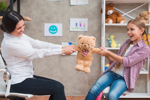 Mujer psicóloga y niña sonriente sentados cara a cara tirando del osito de peluche