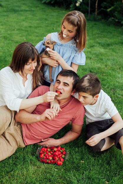 Mujer que alimenta la fresa al marido mientras que se sienta con sus niños en hierba