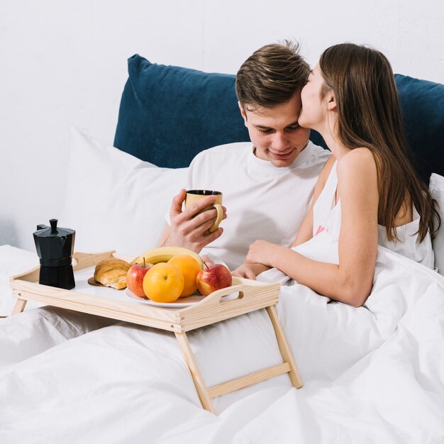 Mujer que besa al hombre en la frente en cama con la bandeja de la comida